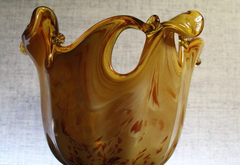 May include: A hand-blown amber glass vase featuring a swirling marbled pattern with deep brown and golden hues. The vessel has an irregular, wavy rim with a prominent cutout opening near the top, creating a fluid and organic silhouette.