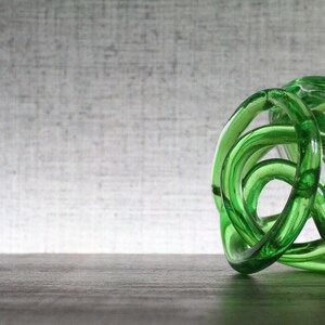 May include: A translucent emerald green glass sculpture formed from a continuous, knotted, and intertwined circular tube. The object features a smooth, polished surface with varying light reflections throughout its complex, rounded, and organic shape.