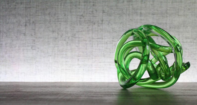 May include: A translucent emerald green glass sculpture formed from a continuous, knotted, and intertwined circular tube. The object features a smooth, polished surface with varying light reflections throughout its complex, rounded, and organic shape.