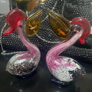 May include: Two glass pelican figurines featuring clear bodies with pink and white marbled patterns, dark blue bases, and amber-colored beaks. Each pelican has a red crest and a small, orange-colored fish encased within the translucent amber beak.