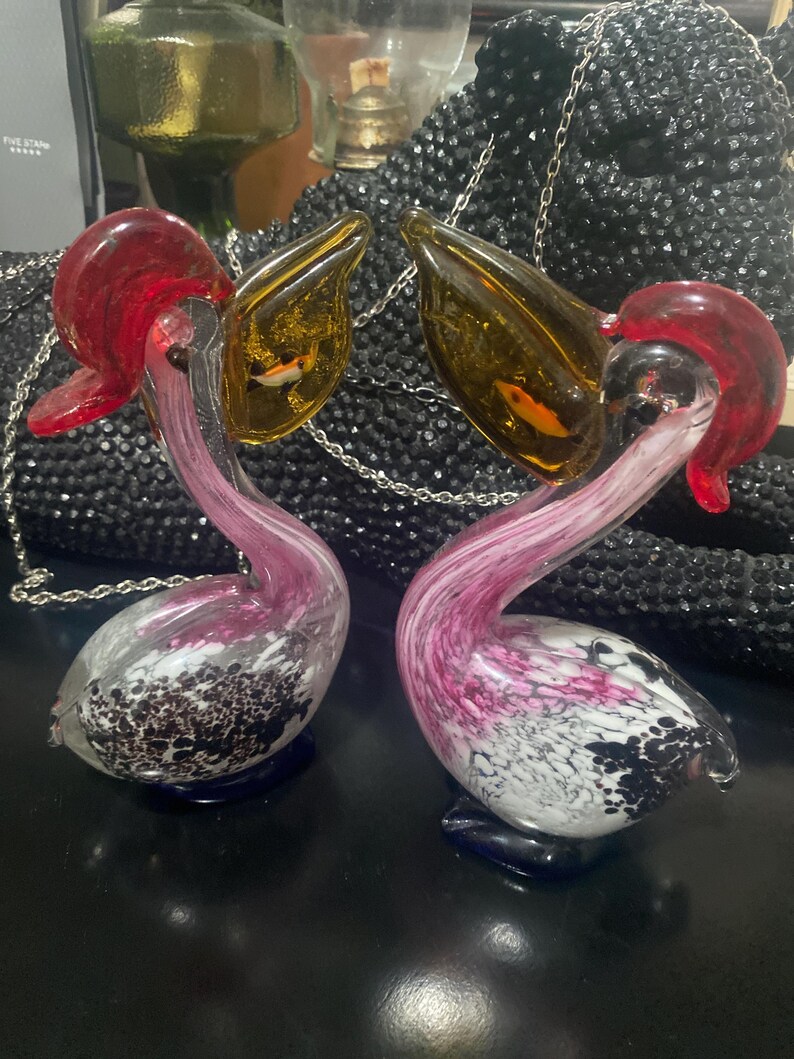 May include: Two glass pelican figurines featuring clear bodies with pink and white marbled patterns, dark blue bases, and amber-colored beaks. Each pelican has a red crest and a small, orange-colored fish encased within the translucent amber beak.