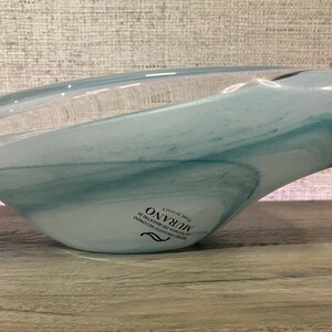 May include: A shallow, asymmetrical glass bowl featuring swirling patterns of translucent white and soft blue. The bowl has a smooth, polished finish and includes a black sticker on the side with the text MURANO MADE IN ITALY.