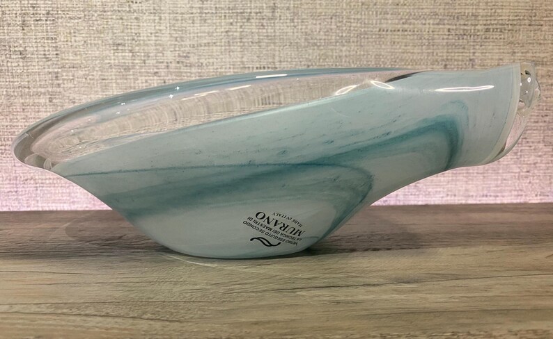 May include: A shallow, asymmetrical glass bowl featuring swirling patterns of translucent white and soft blue. The bowl has a smooth, polished finish and includes a black sticker on the side with the text MURANO MADE IN ITALY.