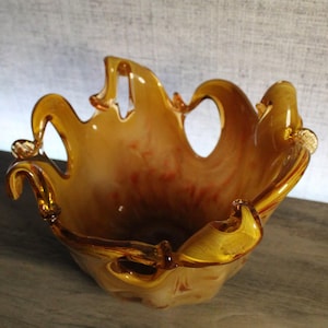 May include: A hand-blown glass bowl featuring a warm amber and cream marbled color palette. The bowl has a distinctively ruffled, open-work rim with several arched cutouts, creating a fluid, organic shape with translucent glass edges.