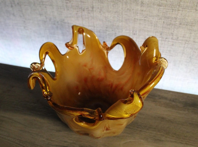 May include: A hand-blown glass bowl featuring a warm amber and cream marbled color palette. The bowl has a distinctively ruffled, open-work rim with several arched cutouts, creating a fluid, organic shape with translucent glass edges.