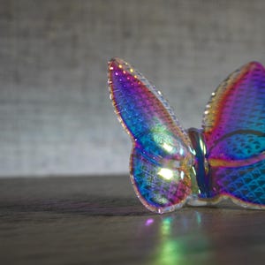 K&ouml;nnte beinhalten: Eine Schmetterlingsfigur aus irisierendem Glas mit einem lebendigen Spektrum aus Blau-, Lila- und Magentat&ouml;nen. Die Fl&uuml;gel weisen ein strukturiertes, geometrisches Schuppenmuster auf, das das Licht einf&auml;ngt und eine reflektierende, bunte Oberfl&auml;che erzeugt.