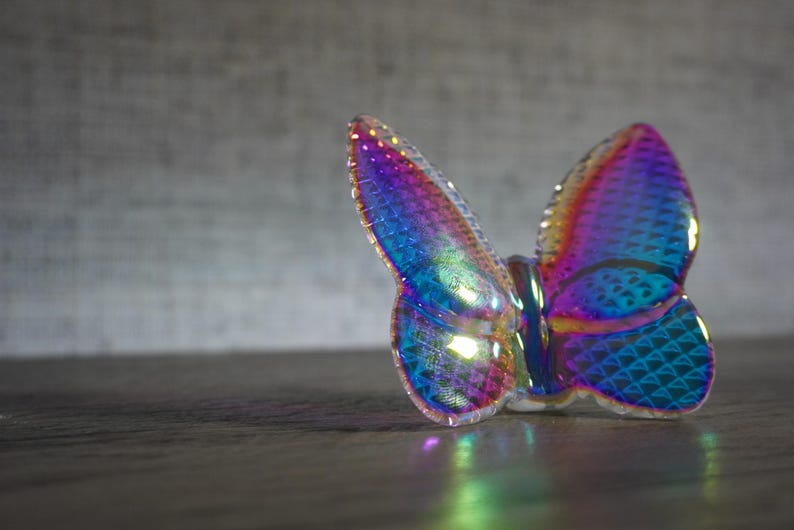 K&ouml;nnte beinhalten: Eine Schmetterlingsfigur aus irisierendem Glas mit einem lebendigen Spektrum aus Blau-, Lila- und Magentat&ouml;nen. Die Fl&uuml;gel weisen ein strukturiertes, geometrisches Schuppenmuster auf, das das Licht einf&auml;ngt und eine reflektierende, bunte Oberfl&auml;che erzeugt.