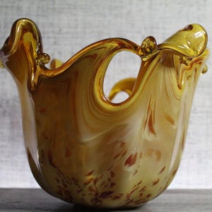 May include: A hand-blown glass bowl featuring a swirling amber and golden-yellow color palette. The vessel has a wide, ruffled, and undulating rim with distinct cut-out sections, creating a fluid, organic shape with translucent glass.