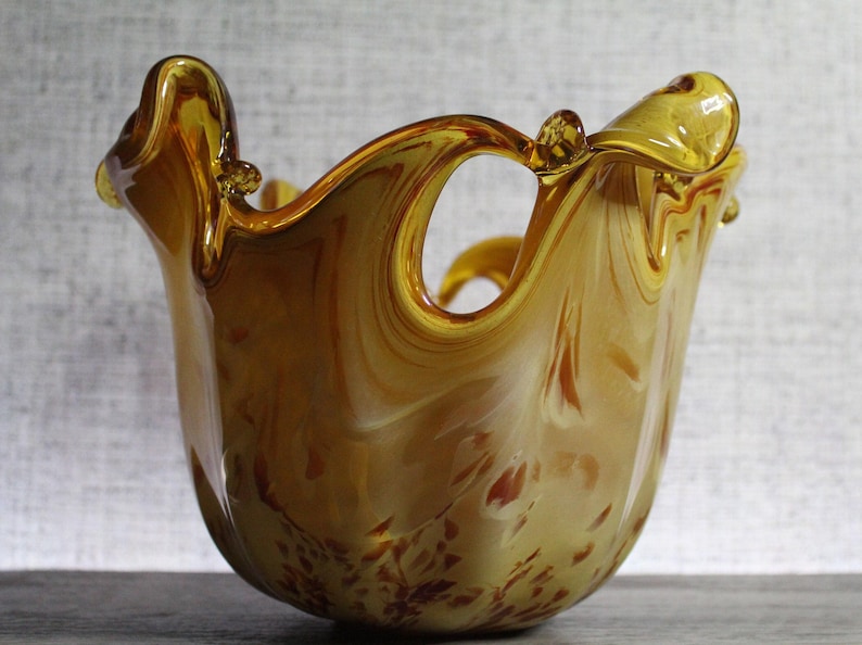 May include: A hand-blown glass bowl featuring a swirling amber and golden-yellow color palette. The vessel has a wide, ruffled, and undulating rim with distinct cut-out sections, creating a fluid, organic shape with translucent glass.