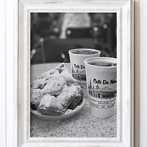May include: Black and white photo of two cups of coffee with the text "Cafe Du Monde" on them. The cups are sitting on a table with a plate of beignets, a type of fried dough pastry.