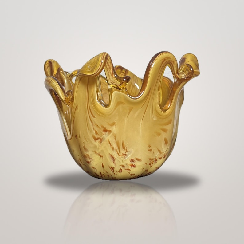 May include: A translucent amber glass bowl with a ruffled, wavy rim. The body of the bowl features a swirling, mottled pattern of golden yellow and darker brown tones, creating a fluid, organic appearance with a smooth, polished finish.