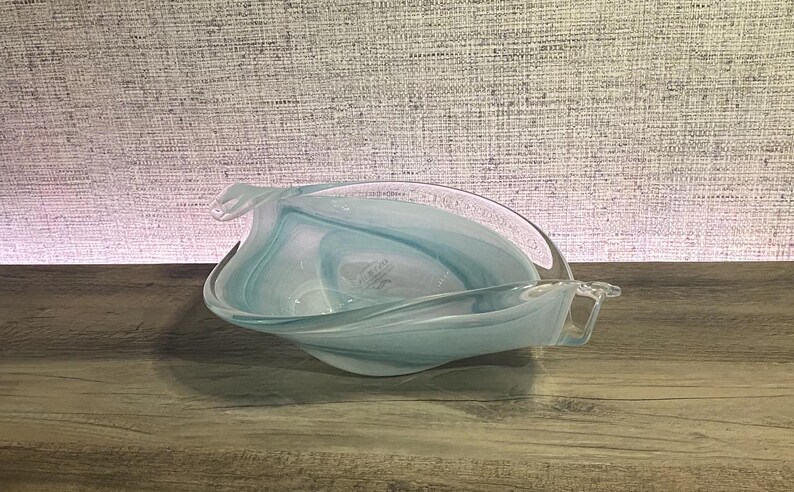 May include: A clear glass bowl featuring swirling patterns of light blue and white. The bowl has an elongated, organic shape with pointed ends and curved, undulating edges that create a fluid, wave-like appearance throughout the glass.