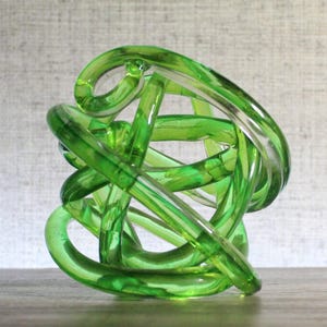 May include: A translucent lime green glass sculpture formed from a continuous, knotted, and intertwined tube. The object features a smooth, polished surface with varying shades of green created by the overlapping layers of the glass tubing.