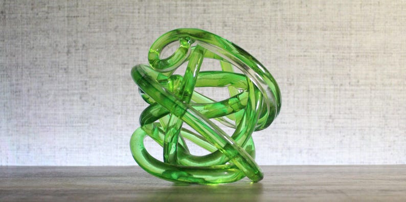 May include: A translucent lime green glass sculpture formed from a continuous, knotted, and intertwined tube. The object features a smooth, polished surface with varying shades of green created by the overlapping layers of the glass tubing.