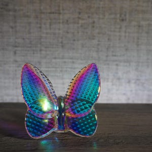 K&ouml;nnte beinhalten: Ein schmetterlingsf&ouml;rmiges Ornament mit einer holografischen, irisierenden Oberfl&auml;che, die zwischen Blau-, Lila- und Gr&uuml;nt&ouml;nen wechselt. Die Oberfl&auml;che weist ein texturiertes, geometrisches Muster auf, das Licht reflektiert und einen bunten Effekt erzeugt.