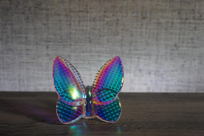 K&ouml;nnte beinhalten: Ein schmetterlingsf&ouml;rmiges Ornament mit einer holografischen, irisierenden Oberfl&auml;che, die zwischen Blau-, Lila- und Gr&uuml;nt&ouml;nen wechselt. Die Oberfl&auml;che weist ein texturiertes, geometrisches Muster auf, das Licht reflektiert und einen bunten Effekt erzeugt.