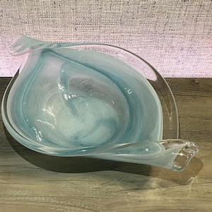May include: A clear glass bowl with a swirling pattern of white and light blue colors. The bowl has an asymmetrical, leaf-like shape with elongated, curved edges that taper to points on opposite sides, creating a fluid and organic appearance.