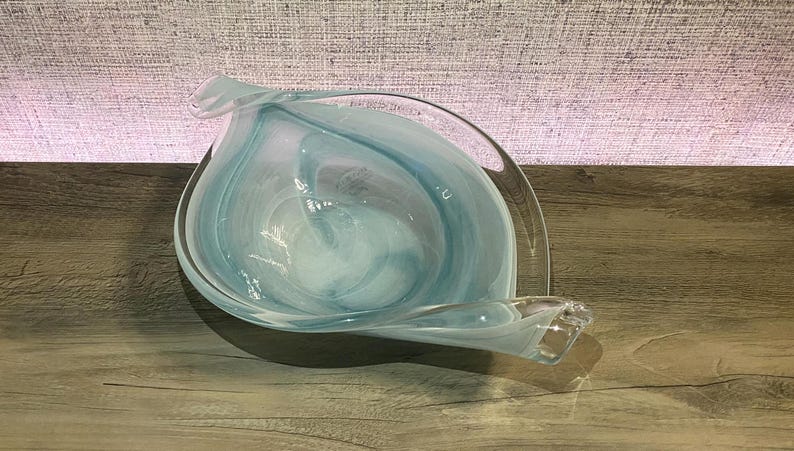 May include: A clear glass bowl with a swirling pattern of white and light blue colors. The bowl has an asymmetrical, leaf-like shape with elongated, curved edges that taper to points on opposite sides, creating a fluid and organic appearance.