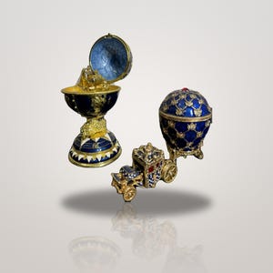 Könnte beinhalten: Eine dekorative, blau-goldene eiförmige Schmuckschatulle mit Kompass-Sockel, die ein kleines goldenes Schiff enthält. Daneben steht eine blau-goldene kutschenförmige Schmuckschatulle mit einem passenden eiförmigen Oberteil.