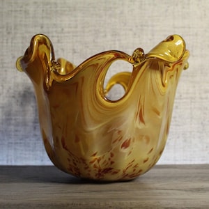 May include: A hand-blown glass bowl featuring a swirling amber and cream color palette. The vessel has a wide, ruffled, and uneven rim with distinct organic curves and a translucent finish that catches the light, creating depth.