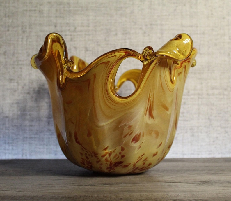 May include: A hand-blown glass bowl featuring a swirling amber and cream color palette. The vessel has a wide, ruffled, and uneven rim with distinct organic curves and a translucent finish that catches the light, creating depth.
