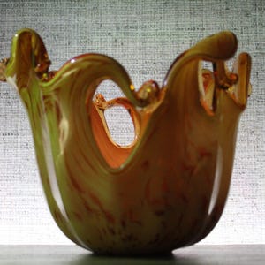 May include: A translucent amber and yellow glass bowl featuring a wide, undulating rim with deep, wave-like cutouts. The glass displays a swirling, marbled color pattern throughout its rounded body, creating a fluid, organic shape.