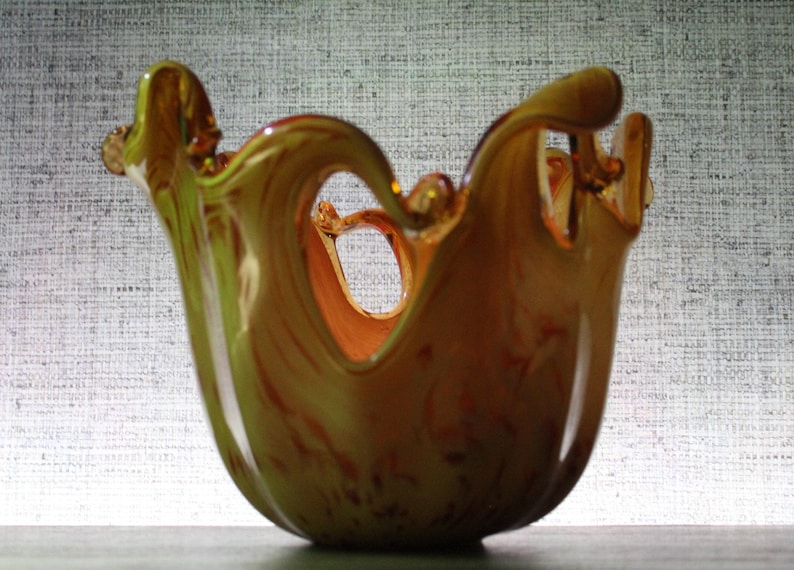 May include: A translucent amber and yellow glass bowl featuring a wide, undulating rim with deep, wave-like cutouts. The glass displays a swirling, marbled color pattern throughout its rounded body, creating a fluid, organic shape.