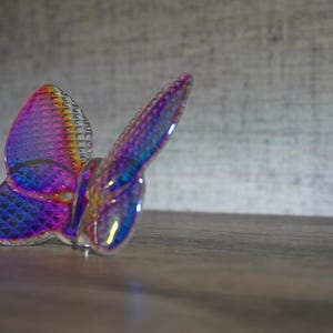 K&ouml;nnte beinhalten: Eine durchscheinende Schmetterlingsfigur mit einer lebendigen, irisierenden Oberfl&auml;che, die zwischen Violett-, Blau- und Oranget&ouml;nen wechselt. Die Fl&uuml;geloberfl&auml;che weist ein markantes, rautenf&ouml;rmiges Gittermuster auf, das das Licht reflektiert.