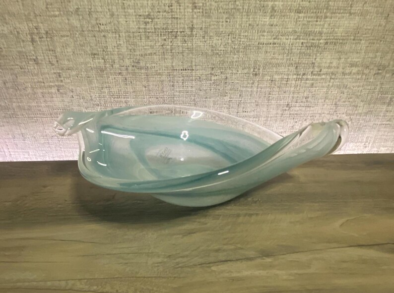 May include: A decorative glass bowl featuring a fluid, elongated leaf-like shape with swirling patterns of translucent light blue and white colors. The bowl has smooth, curved edges and a polished finish, creating a soft, layered appearance.