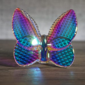 K&ouml;nnte beinhalten: Eine durchscheinende Schmetterlingsfigur mit einer lebendigen, irisierenden Oberfl&auml;che, die zwischen Violett-, Blau- und Gr&uuml;nt&ouml;nen wechselt. Die Fl&uuml;gel zeigen ein sich wiederholendes geometrisches Gittermuster, das das Licht einf&auml;ngt.