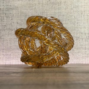 May include: A glass sculpture in the shape of a knot. The sculpture is made of clear glass with a golden amber hue.