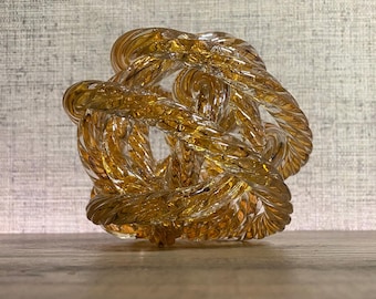 May include: A glass sculpture in the shape of a knot. The sculpture is made of clear glass with a golden amber hue.