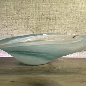 May include: A hand-blown glass bowl featuring an elongated, asymmetrical shape with a smooth, polished surface. The glass displays a swirling blend of translucent white, soft teal, and pale blue hues, creating a marbled effect throughout the piece.