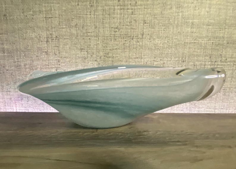 May include: A hand-blown glass bowl featuring an elongated, asymmetrical shape with a smooth, polished surface. The glass displays a swirling blend of translucent white, soft teal, and pale blue hues, creating a marbled effect throughout the piece.