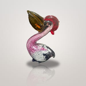 May include: A glass pelican figurine featuring a translucent amber beak, a vibrant red crest, and a curved neck with swirling pink and white patterns. The body displays a mottled black and white texture, capturing the bird in a standing pose.