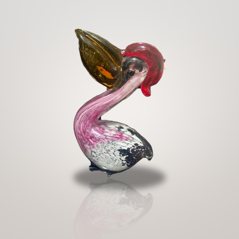 May include: A glass pelican figurine featuring a translucent amber beak, a vibrant red crest, and a curved neck with swirling pink and white patterns. The body displays a mottled black and white texture, capturing the bird in a standing pose.