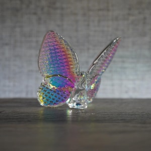 K&ouml;nnte beinhalten: Eine Schmetterlingsfigur aus klarem Glas mit einer irisierenden, regenbogenfarbenen Oberfl&auml;che auf den Fl&uuml;geln. Die Fl&uuml;gel weisen ein strukturiertes Gittermuster auf, das das Licht einf&auml;ngt und schimmernde Blau-, Rosa- und Goldt&ouml;ne erzeugt.