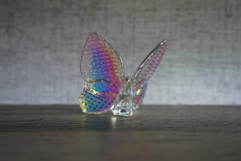 K&ouml;nnte beinhalten: Eine Schmetterlingsfigur aus klarem Glas mit einer irisierenden, regenbogenfarbenen Oberfl&auml;che auf den Fl&uuml;geln. Die Fl&uuml;gel weisen ein strukturiertes Gittermuster auf, das das Licht einf&auml;ngt und schimmernde Blau-, Rosa- und Goldt&ouml;ne erzeugt.
