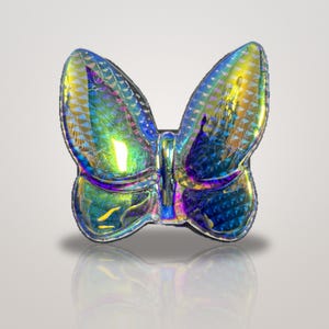 K&ouml;nnte beinhalten: Ein schmetterlingsf&ouml;rmiges Objekt mit einer stark reflektierenden, irisierenden Oberfl&auml;che, die zwischen Blau-, Gr&uuml;n- und Goldt&ouml;nen wechselt. Die Fl&uuml;gel weisen ein strukturiertes, geometrisches Muster auf, das das Licht einf&auml;ngt.