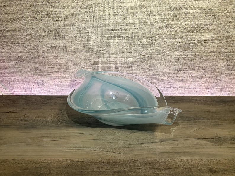 May include: A decorative glass bowl featuring a swirling pattern of translucent light blue and opaque white colors. The bowl has an elongated, asymmetrical leaf-like shape with curved, undulating edges that taper to pointed ends on both sides.