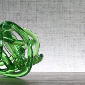 May include: A translucent emerald green glass sculpture formed from a continuous, knotted, and intertwined tube. The object features a smooth, polished surface with varying light reflections throughout its complex, rounded, and organic shape.