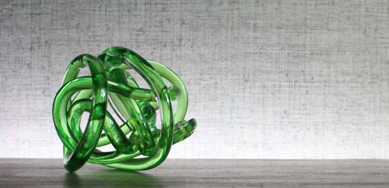 May include: A translucent emerald green glass sculpture formed from a continuous, knotted, and intertwined tube. The object features a smooth, polished surface with varying light reflections throughout its complex, rounded, and organic shape.