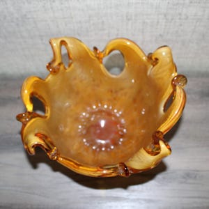 May include: A hand-blown glass bowl featuring a warm amber and golden-brown color palette with a mottled, translucent finish. The bowl has a distinctively ruffled, organic rim with several open loops and a deep, rounded base.