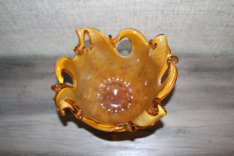 May include: A hand-blown glass bowl featuring a warm amber and golden-brown color palette with a mottled, translucent finish. The bowl has a distinctively ruffled, organic rim with several open loops and a deep, rounded base.