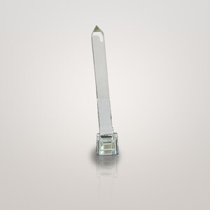 May include: A clear, transparent glass obelisk standing upright on a square, faceted glass base. The tall, slender, four-sided column tapers to a pointed pyramid top, reflecting light through its smooth, polished surfaces and edges.