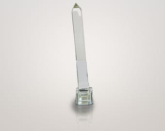 May include: A clear, transparent glass obelisk standing upright on a square, faceted glass base. The tall, slender, four-sided column tapers to a pointed pyramid top, reflecting light through its smooth, polished surfaces and edges.