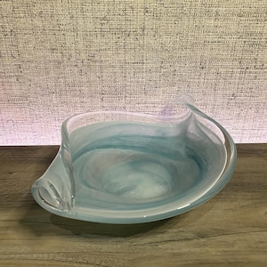 May include: A shallow glass bowl featuring a swirling pattern of light blue and white colors. The bowl has an asymmetrical, organic shape with curved, elevated edges that taper into points on opposite sides, creating a fluid appearance.
