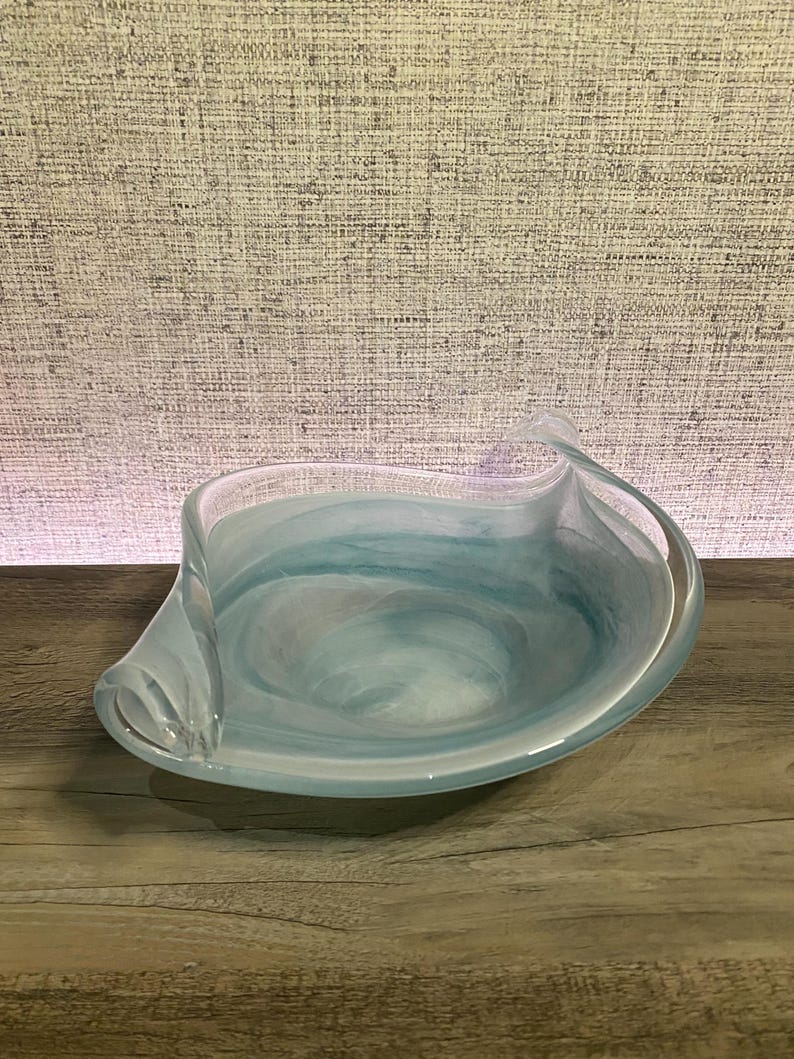 May include: A shallow glass bowl featuring a swirling pattern of light blue and white colors. The bowl has an asymmetrical, organic shape with curved, elevated edges that taper into points on opposite sides, creating a fluid appearance.
