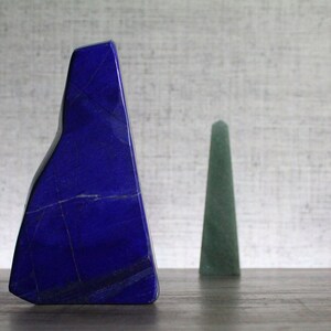 Op de afbeelding: Een groot, blauw, onregelmatig gevormd lapis lazuli-kristal naast een kleiner, groen, puntig kristal. Beide kristallen liggen op een licht houten oppervlak.