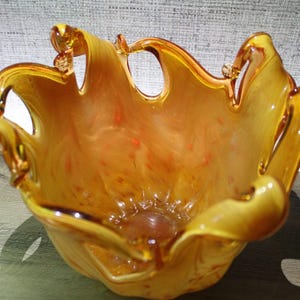 May include: A hand-blown glass bowl featuring a vibrant amber and orange swirl pattern. The bowl has a wide, organic, ruffled rim with several distinct, curved glass loops extending upward, creating a dynamic, open, and fluid silhouette.